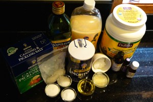 An assortment of ingredients for a creamy hand scrub: olive oil, hand soap, coconut oil, salts, essential oils, and jojoba oil.